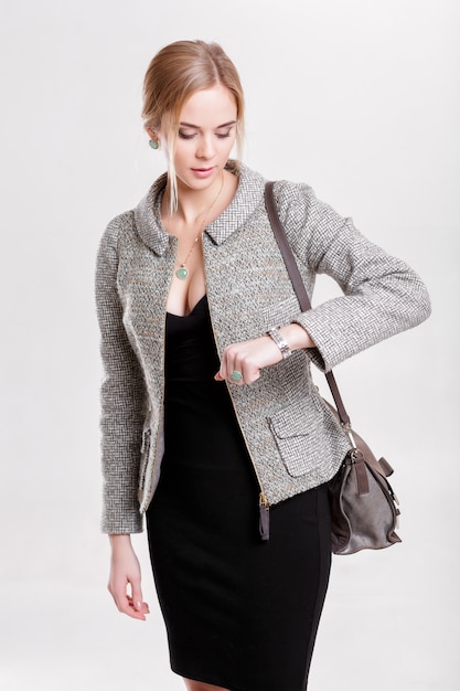 Young beautiful business woman blonde in black dress, jacket and purse looks at her watch on gray background. girl is late on time