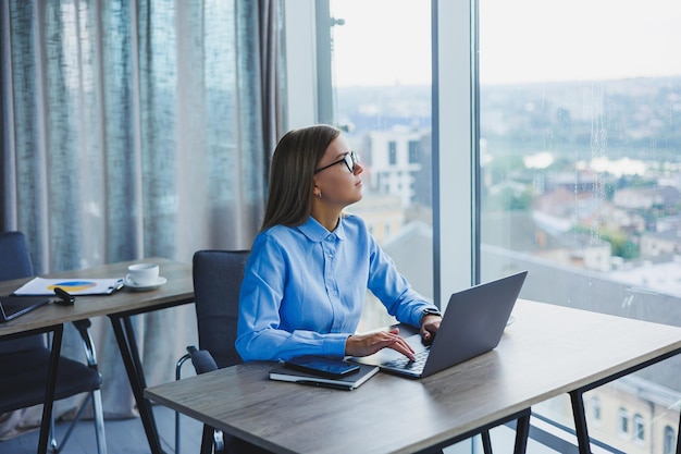 Young beautiful business woman in a blue shirt sitting in the office with a laptop and doing business