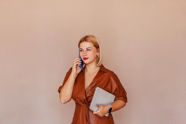 그녀의 손에 노트북으로 전화 통화 하는 갈색 드레스에 젊은 아름 다운 사업가