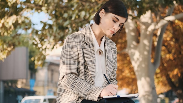 Young beautiful businesswoman sitting on cozy street and drawing up business plan for a month in her notepad