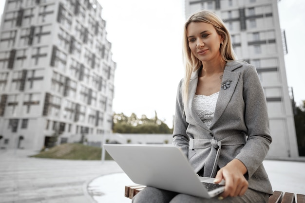 Young beautiful businesswoman working on laptop sitting on the bench in the street