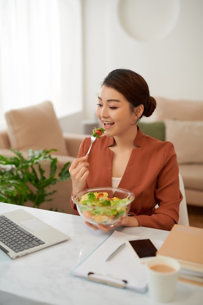 Young beautiful businesswoman at working place, eating vegetable salad in bowl,