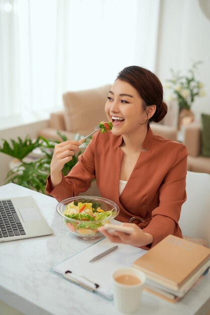 Photo young beautiful businesswoman at working place, eating vegetable salad in bowl,