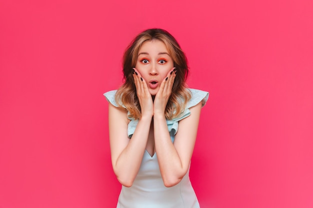 A young beautiful caucasian impressed brownhaired woman with bright make up in a stylish light blue dress surprised by the news about a sale and discounts  isolated on a bright color pink wall