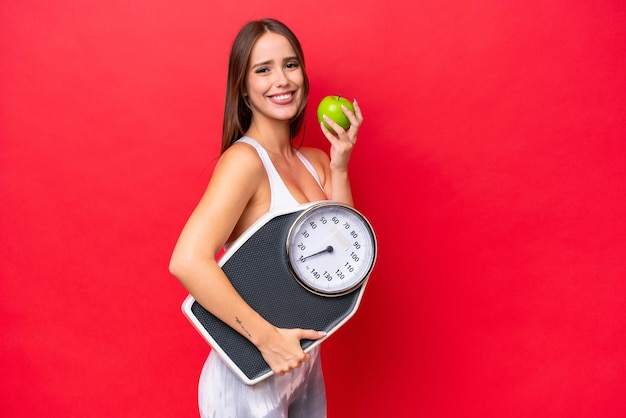 Young beautiful caucasian woman isolated on red background with weighing machine and with an apple