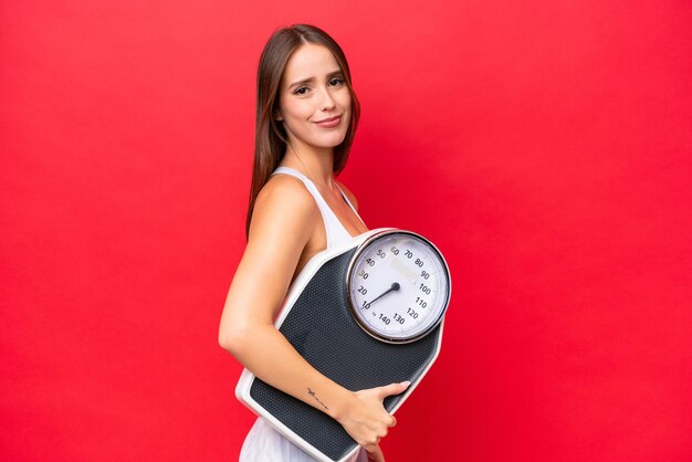 Young beautiful caucasian woman isolated on red background with weighing machine