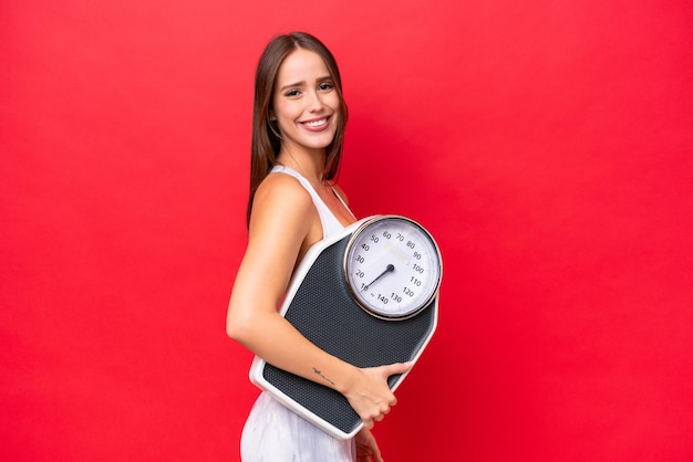 Young beautiful caucasian woman isolated on red background with weighing machine