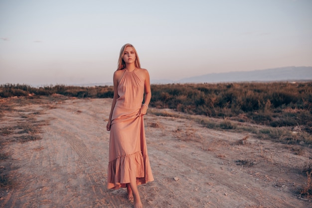 Young beautiful Caucasian woman in pink dress walks on sand with bare feet during sunset