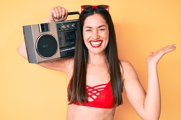 Young beautiful caucasian woman wearing bikini and holding boombox celebrating victory with happy smile and winner expression with raised hands