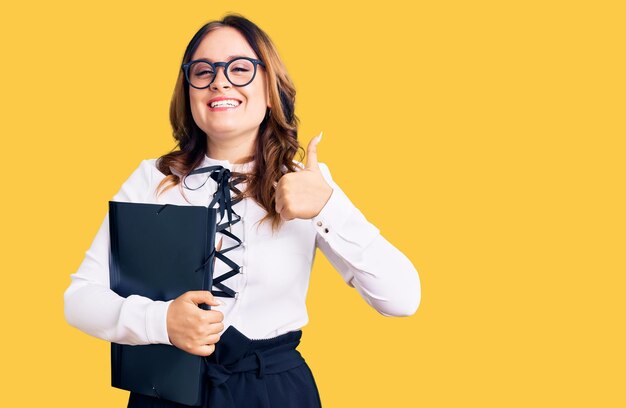 Young beautiful caucasian woman wearing business shirt and glasses holding folder smiling happy and positive thumb up doing excellent and approval sign