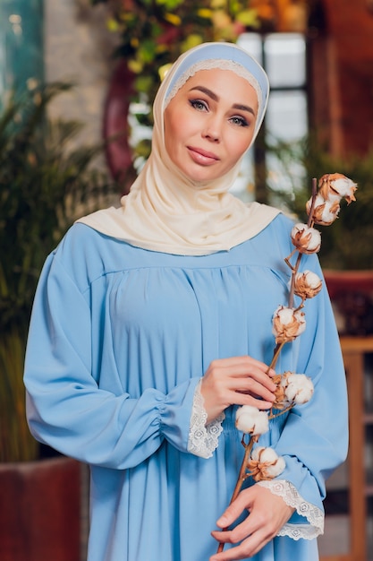 大きなフルレングスの窓のある流行に敏感なコーヒーショップで伝統的なイスラム教徒のスカーフを身に着けている若い美しい白人女性。居心地の良いカフェで青いヒジャーブの女性。背景、コピースペース、ポートレートをクローズアップ。