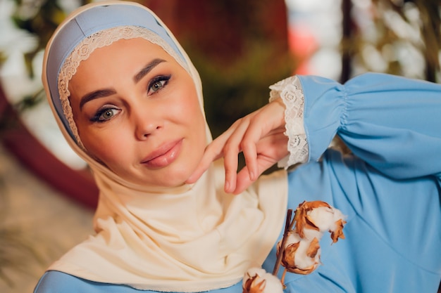 큰 전체 길이 창문이 힙 스터 커피 숍에서 전통적인 이슬람 Headscarf를 입고 젊은 아름 다운 백인 여자. 아늑한 카페에서 파란색 Hijab의 여성. 배경, 복사 공간, 초상화를 닫습니다.
