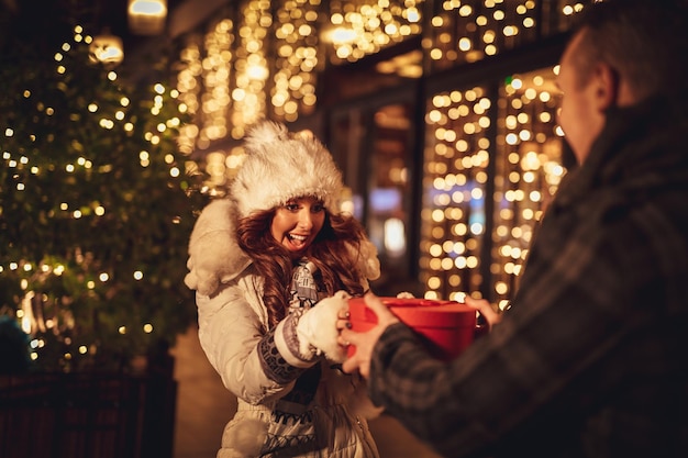 街の通りでクリスマスを祝い、お互いに贈り物を与える若い美しい陽気なカップル。