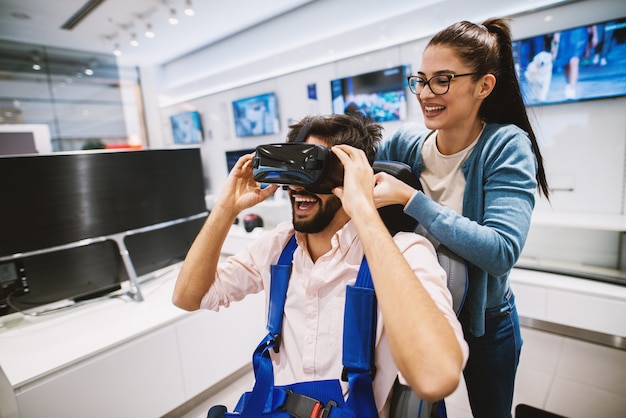 Young beautiful cheerful girl is helping a young joyful man setup VR system on him and tie the security belt of a chair he is sitting on.