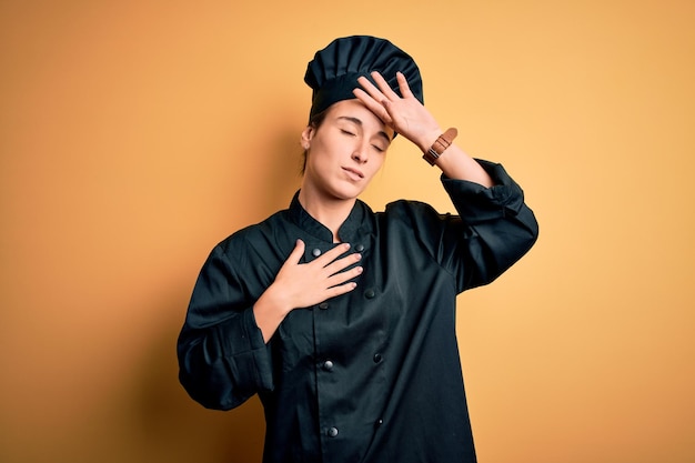 Young beautiful chef woman wearing cooker uniform and hat standing over yellow background Touching forehead for illness and fever flu and cold virus sick