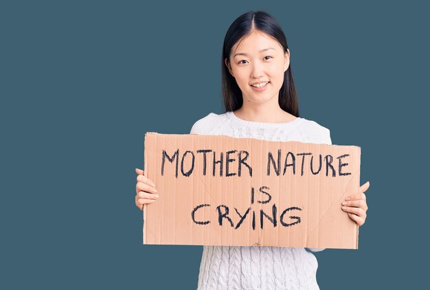 Young beautiful chinese woman holding mother nature is crying cardboard banner looking positive and happy standing and smiling with a confident smile showing teeth