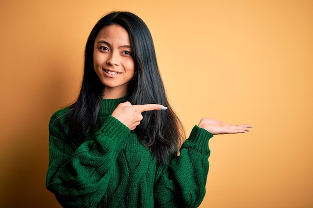 Молодая красивая китаянка в зеленом свитере на изолированном желтом фоне удивлена и улыбается в камеру, показывая рукой и указывая пальцем