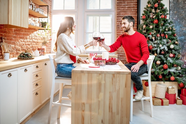 Young beautiful couple dressed in sweaters celebrating New Year drinking wine in the apartment with Christmas tree