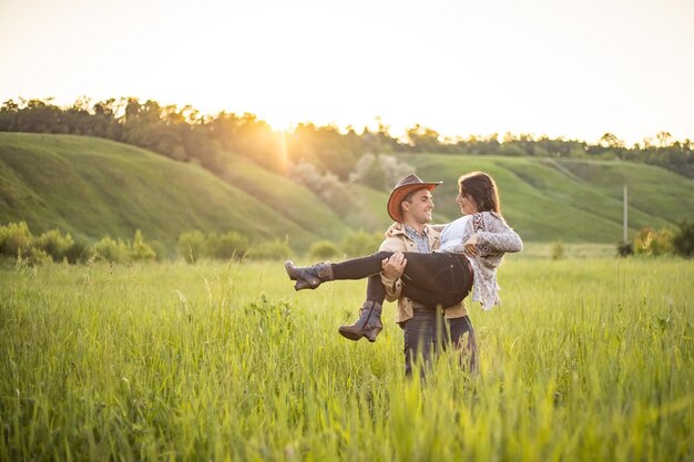 夕日の光線で緑の牧草地に若い美しいカップル男は彼の腕のカウボーイ服素朴なスタイルで女の子を保持します