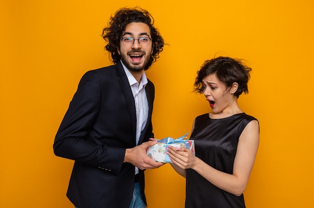 Young beautiful couple happy man giving a present to his surprised and amazed girlfriend celebrating international women's day march 8 standing over orange background