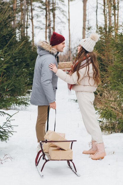 Young beautiful couple hugs and kisses in a winter coniferous forest and carries boxes with gifts on a sled. A park with Christmas trees on the background. Christmas mood. Tinting.