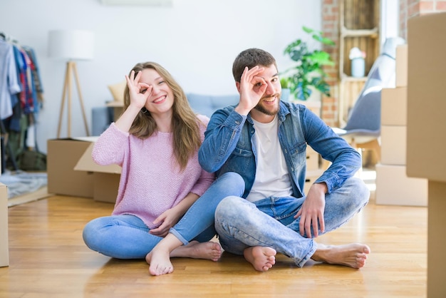 Young beautiful couple moving to a new house sitting on the floor doing ok gesture with hand smiling eye looking through fingers with happy face