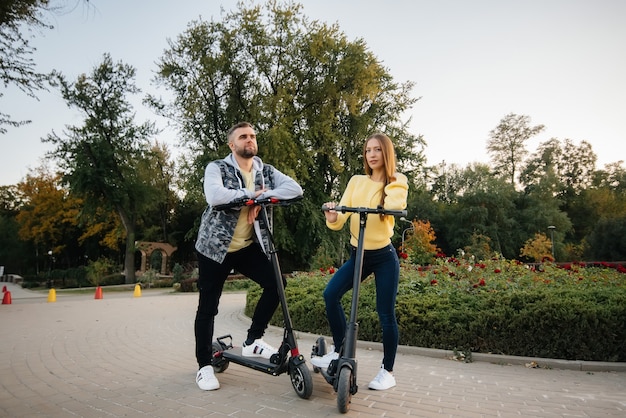 若い美しいカップルが暖かい秋の日に公園で電動スクーターに乗る