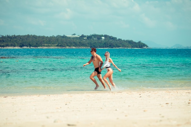 Young beautiful couple runs on the beach hand in hand, the husband in swim trousers and his lovely wife with long white cornrows and white one-piece swimsuit; wide shot.