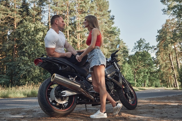 Young beautiful couple talking and having fun sitting on a motorcycle traveling together on a forest