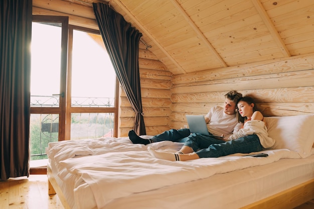 Young beautiful couple watching a movie on a laptop while lying on a bed in a wooden apartment