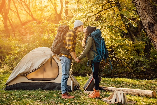 Young beautiful couple with hiking backpack kissing in the forest near tent