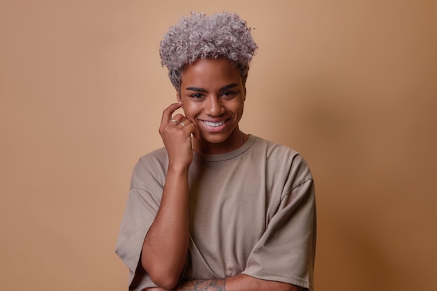 Young beautiful curly African American woman looking at you cutely smiling and touching face wearing beige casual tshirt posing on brown studio background People emotions lifestyle