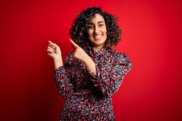 Young beautiful curly arab woman wearing casual floral dress standing over red background smiling and looking at the camera pointing with two hands and fingers to the side