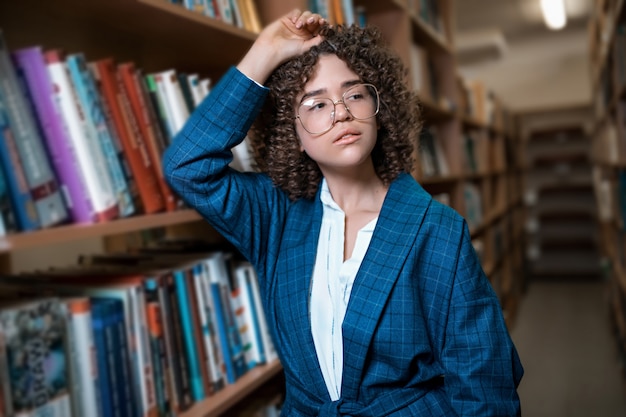 Young beautiful curly girl in glasses and a blue suit is standing in the library