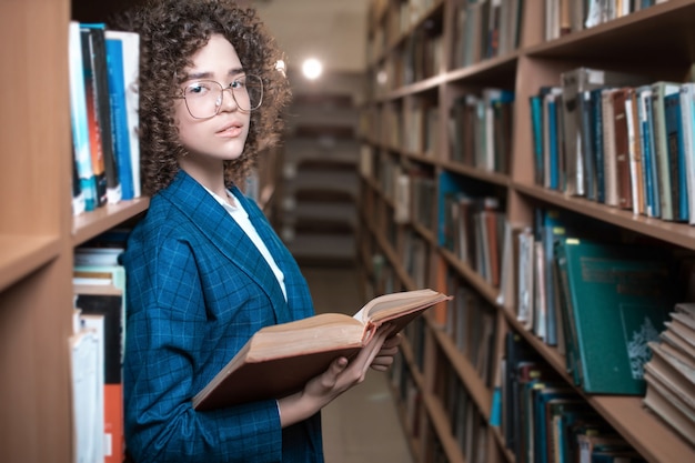 Young beautiful curly girl in glasses and a blue suit is standing in the library