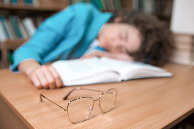 Young beautiful curly girl in glasses and blue suit sitting with books in the library.