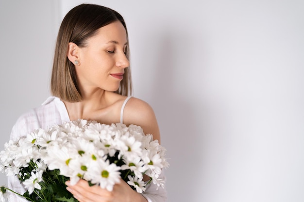 写真 白い壁の背景に白い新鮮な花の花束を保持している若い美しいかわいい甘い素敵な笑顔の女性