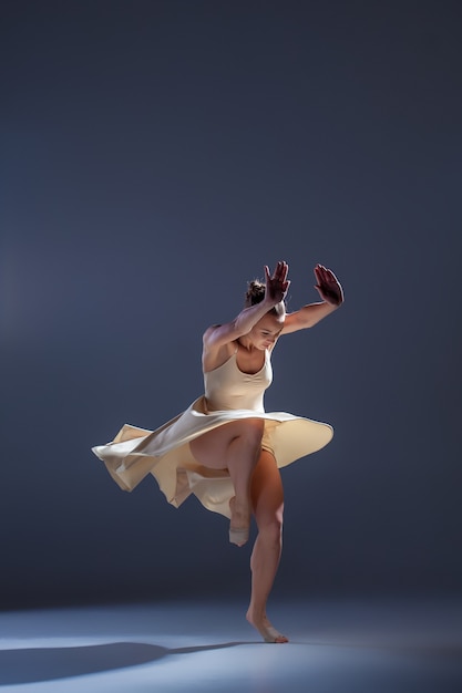 Молодая красивая танцовщица в бежевом платье танцует на сером фоне