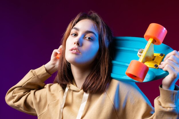 パーカーに身を包んだ若い美しいファッショナブルなヒップスターの女の子は、紫色のスタジオの背景にスケートボードを手に持っていますロリポップを食べます