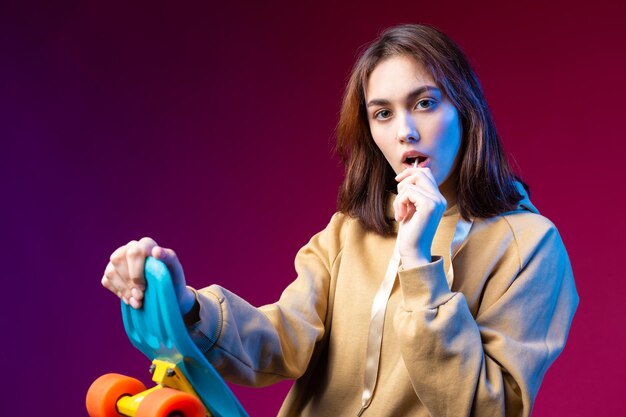 Young beautiful fashionable hipster girl dressed in a hoodie holds a skateboard in her hands on a purple studio background Eats a lollipop