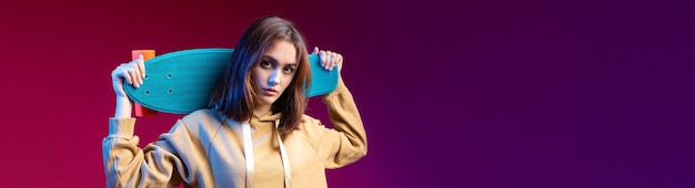 Young beautiful fashionable hipster girl dressed in a hoodie holds a skateboard in her hands on a purple studio background