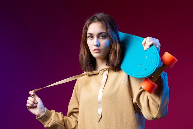 후드티를 입은 젊고 세련된 힙스터 소녀는 보라색 스튜디오 배경에서 스케이트보드를 손에 들고 있다