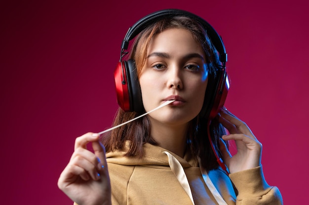 スタジオの赤い劇的な背景で赤いヘッドフォンで音楽を聴いているパーカーに身を包んだ若い美しいファッショナブルなヒップスターの女の子