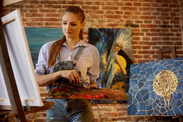 Young beautiful female artist painting on canvas using oil paintings and palette knife painter creat...