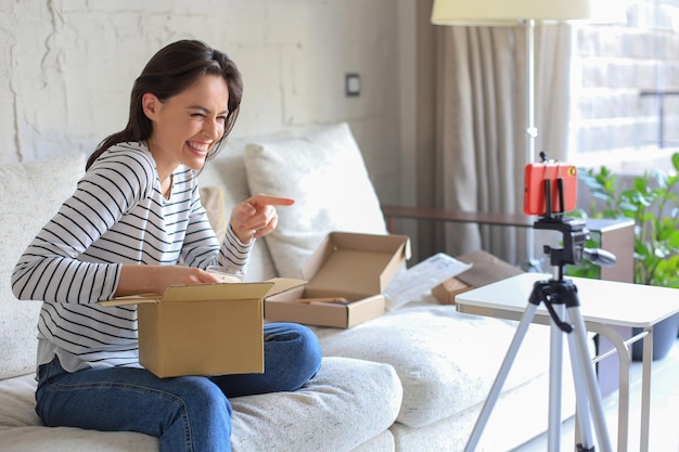 Young beautiful female blogger recording video while unpacking parcel at home