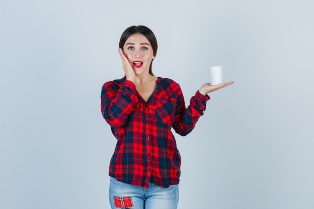 Young beautiful female in casual shirt, jeans holding plastic glass, keeping hand on face and looking puzzled , front view.