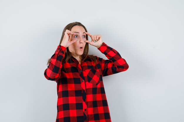 Young beautiful female in casual shirt looking through fingers and looking playful , front view.