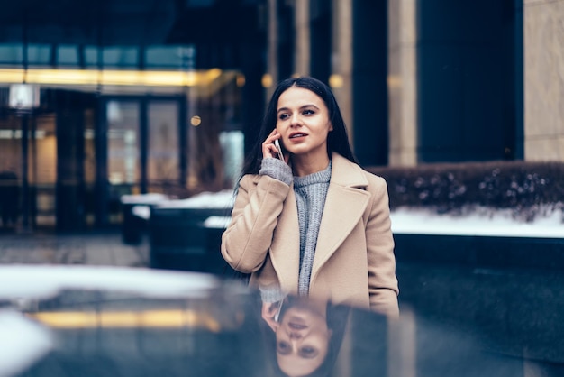 Young beautiful female in coat standing while talking on smartphone