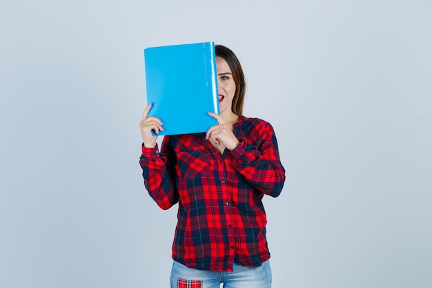 Young beautiful female covering eye with folder in casual shirt, jeans and looking curious. front view.