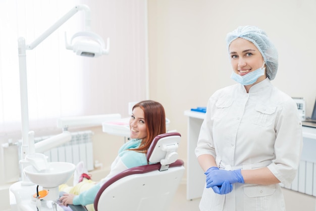 若い美しい女性歯科医と幸せな患者は、カメラを直接見つめています。赤毛の女性は矯正医と一緒に椅子に座り、笑顔で歯科サービスを行っています